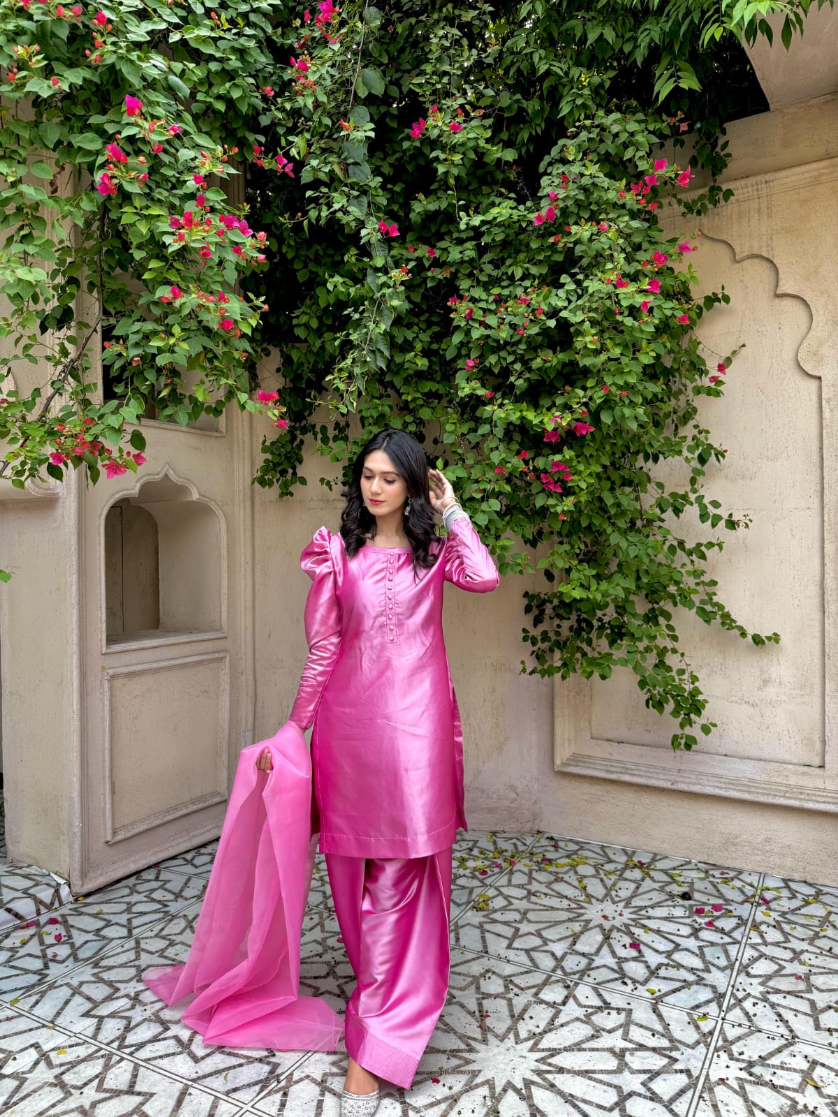 Full standing view of Rose Pink China Katan farshi suit with puff sleeves, wide farshi trouser and organza dupatta beside a floral garden wall
